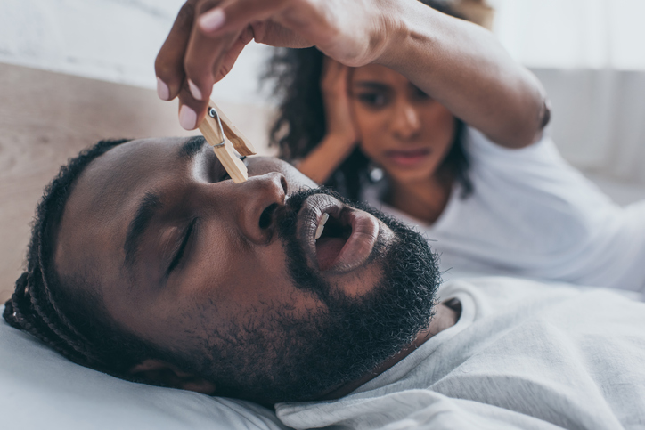 Man asleep in bed snoring, woman holding wooden clip over his nose