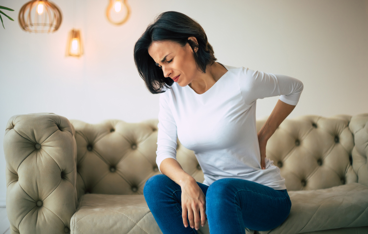 Woman sitting on a couch and holding her lower back with her left hand and looking in pain
